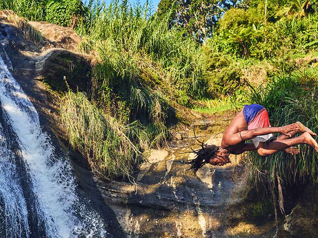 A man somersaults into water with a waterfall in the background