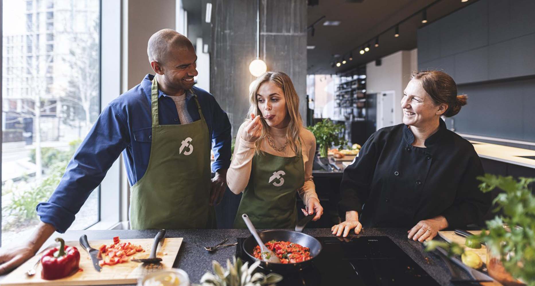 A woman tastes the dish she is cooking at a cooking class, whilst her partner and a chef look on
